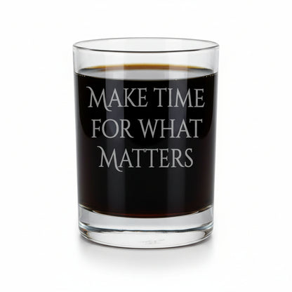 A clear rocks glass with the phrase 'MAKE TIME FOR WHAT MATTERS' etched on the front, filled with a dark amber whiskey, set on a light wood surface against a white background.
