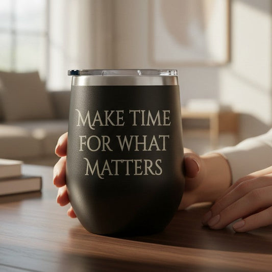 A black insulated wine tumbler with a clear lid sits on a wooden table in a softly lit living room. A person's hands are holding the tumbler, which has the phrase 'MAKE TIME FOR WHAT MATTERS' printed in white serif text.