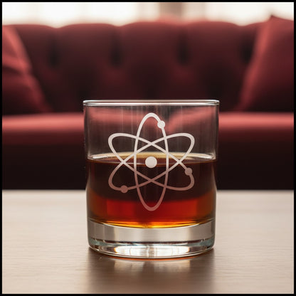 A clear rocks glass with an etched white atom symbol, filled with an amber-brown liquid, sitting on a wooden table with a blurred red sofa in the background.