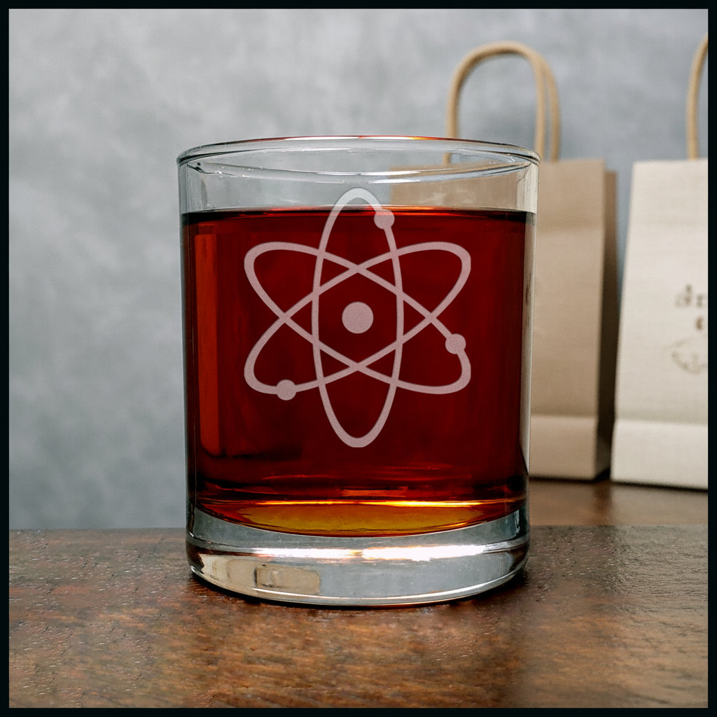 A clear rocks glass filled with amber liquid, featuring a white etched atom symbol on the front, set on a wooden surface 