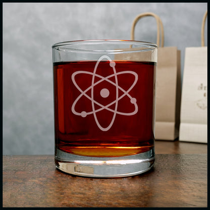 A clear rocks glass filled with amber liquid, featuring a white etched atom symbol on the front, set on a wooden surface 