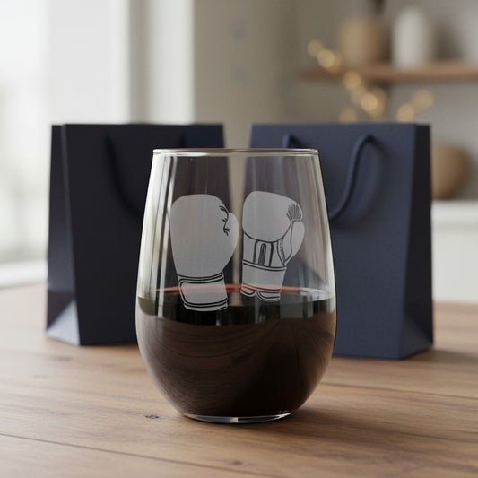 A clear stemless wine glass with an etched boxing motif, partially filled with red wine, set on a wooden table with two dark navy gift bags in the background.