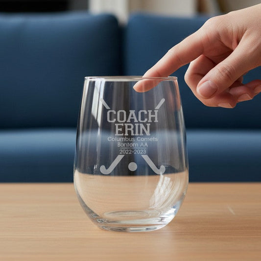 A clear stemless wine glass with white engraved text reading 'COACH ERIN' and crossed golf clubs, placed on a wooden table with a hand pointing toward it. A blue sofa and blurred indoor background are visible.