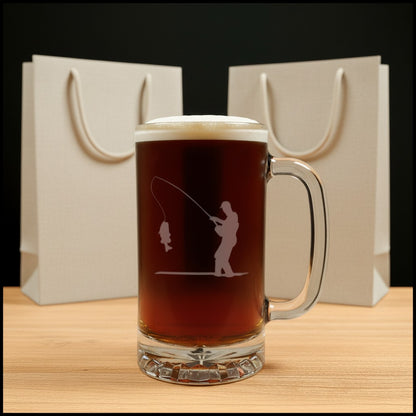 A clear glass beer mug filled with dark amber beer, featuring an etched design of a fisherman casting a line. The mug sits on a light wood surface with two blank beige gift bags against a black background.