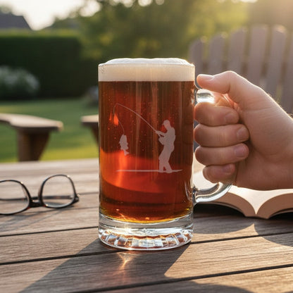 A hand holds a frosted glass beer mug filled with amber beer, featuring an etched fishing silhouette, on a wooden outdoor table with glasses and an open book nearby.