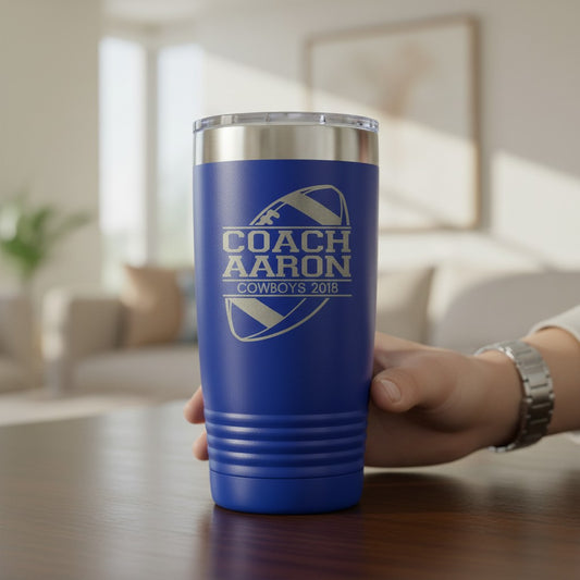 A blue insulated tumbler with a clear lid and silver rim, held by a hand on a wooden table. The tumbler features a laser-engraved design that reads 'COACH AARON COWBOYS 2018' with a football graphic.