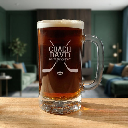 A clear glass beer mug with a handle, filled with dark beer and topped with foam, sitting on a wooden table in a blurred living room setting. The mug is personalized with the etched text 'COACH DAVID Cordoba Crushers 2022-2023' and a hockey design.