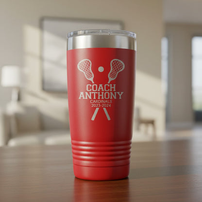 A red insulated tumbler with a clear lid sits on a wooden table in a blurred indoor room. The tumbler is laser engraved with white lacrosse-themed graphics and the text 'COACH ANTHONY CARDINALS 2023-2024'.