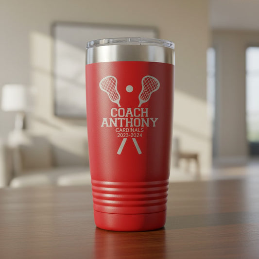 A red insulated tumbler with a clear lid sits on a wooden table in a blurred indoor room. The tumbler is laser engraved with white lacrosse-themed graphics and the text 'COACH ANTHONY CARDINALS 2023-2024'.