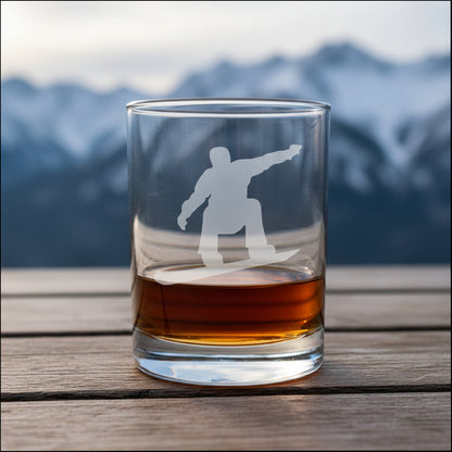 Clear glass tumbler with a snowboarder silhouette etched on it, filled with amber liquid on a wooden surface.