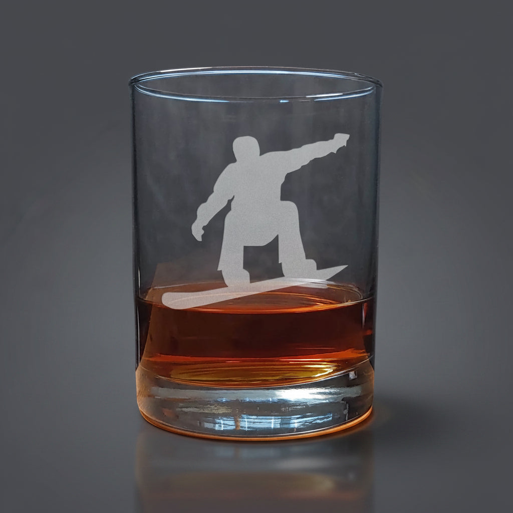 Clear glass tumbler with a snowboarder silhouette etched on it, filled with amber liquid on a wooden surface.