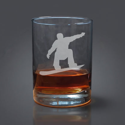 Clear glass tumbler with a snowboarder silhouette etched on it, filled with amber liquid on a wooden surface.