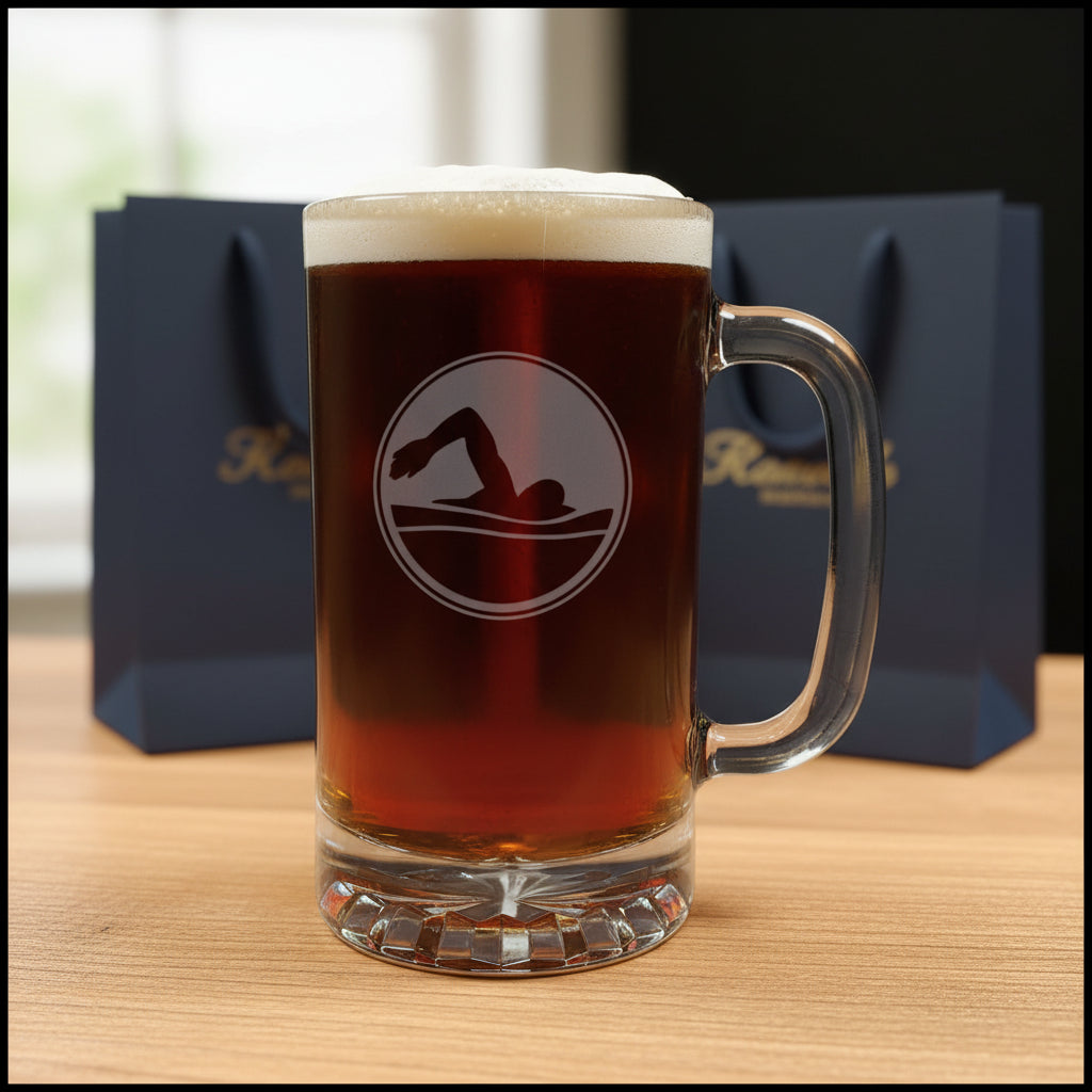 A clear glass beer mug filled with dark amber beer and topped with white foam, standing on a light wood surface against a black background. The mug has a etched circular design with a dark swimmer silhouette.