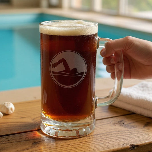 A hand holds the handle of a large clear glass beer mug filled with amber beer, featuring an etched circular image with a swimmer silhouette, set on a wooden table next to a swimming pool.