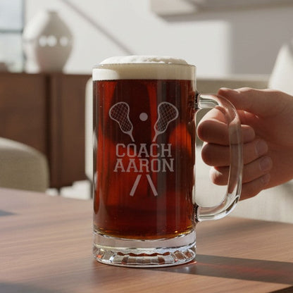 A hand holds a frosted glass mug filled with an amber drink on a wooden table in a bright living room. The mug is etched with a lacrosse graphic and the text 'COACH AARON'.