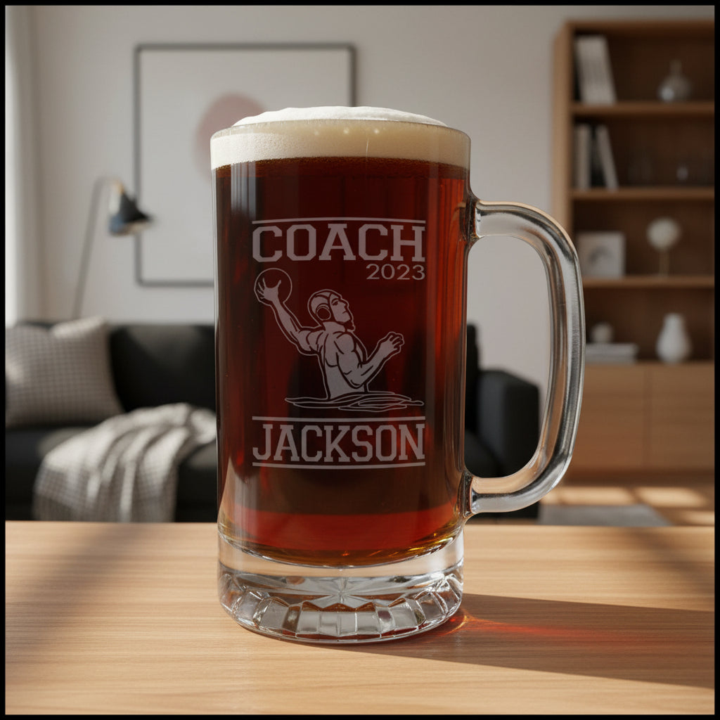 A clear glass beer mug with a handle, filled with dark amber beer and topped with white foam, standing on a light wood surface. The mug is etched with the words 'COACH 2023' and 'JACKSON' around a basketball player graphic.