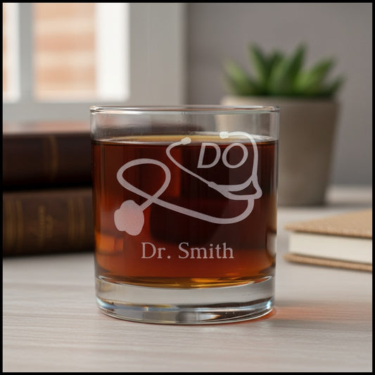A clear glass rocks tumbler filled with amber liquid, etched with a stethoscope and the words 'DO' and 'Dr. Smith', set on a tabletop with blurred books and a plant in the background.