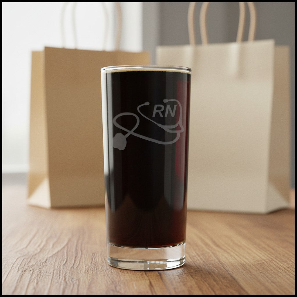 A clear highball glass filled with black coffee, featuring a gray etched stethoscope and 'RN' design, set on a wooden table with blurred paper bags in the background.