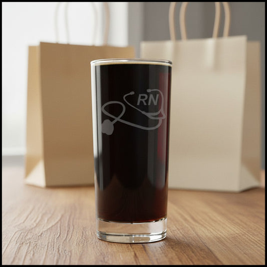 A clear highball glass filled with black coffee, featuring a gray etched stethoscope and 'RN' design, set on a wooden table with blurred paper bags in the background.