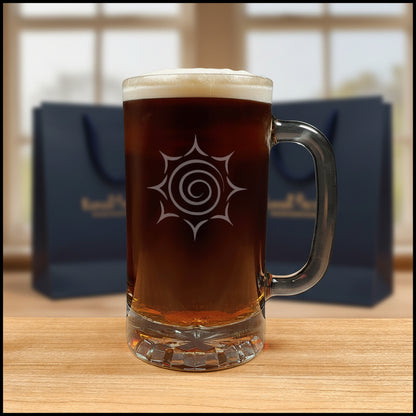 A clear glass beer mug with a handle, filled with dark amber beer and topped with white foam, featuring a etched spiral sunburst design on the front.