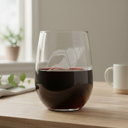 A clear stemless wine glass with an etched seashell design, partially filled with red wine, sitting on a light wooden table in a softly lit room.