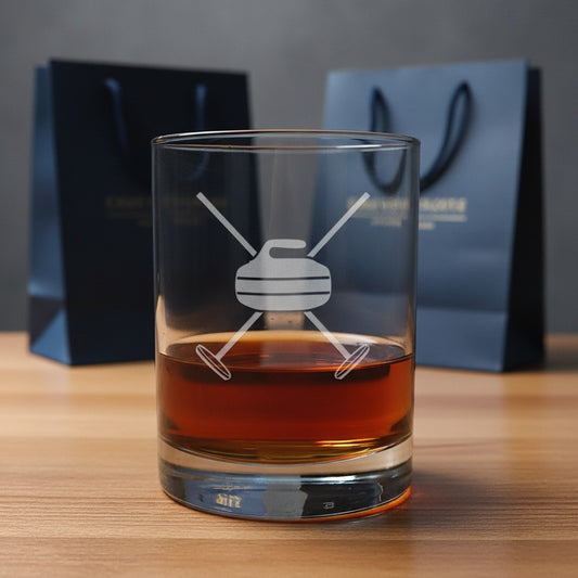 A clear glass rocks tumbler with an etched curling design, filled with amber whiskey, set on a wooden surface with dark blue gift bags in the background.