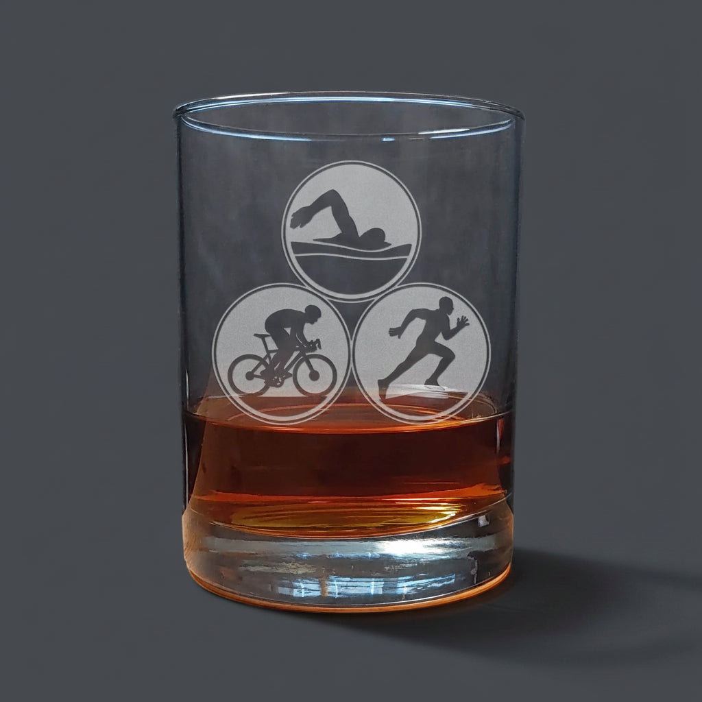 Clear glass tumbler with Triathlon symbols filled with amber liquid on a wooden surface.