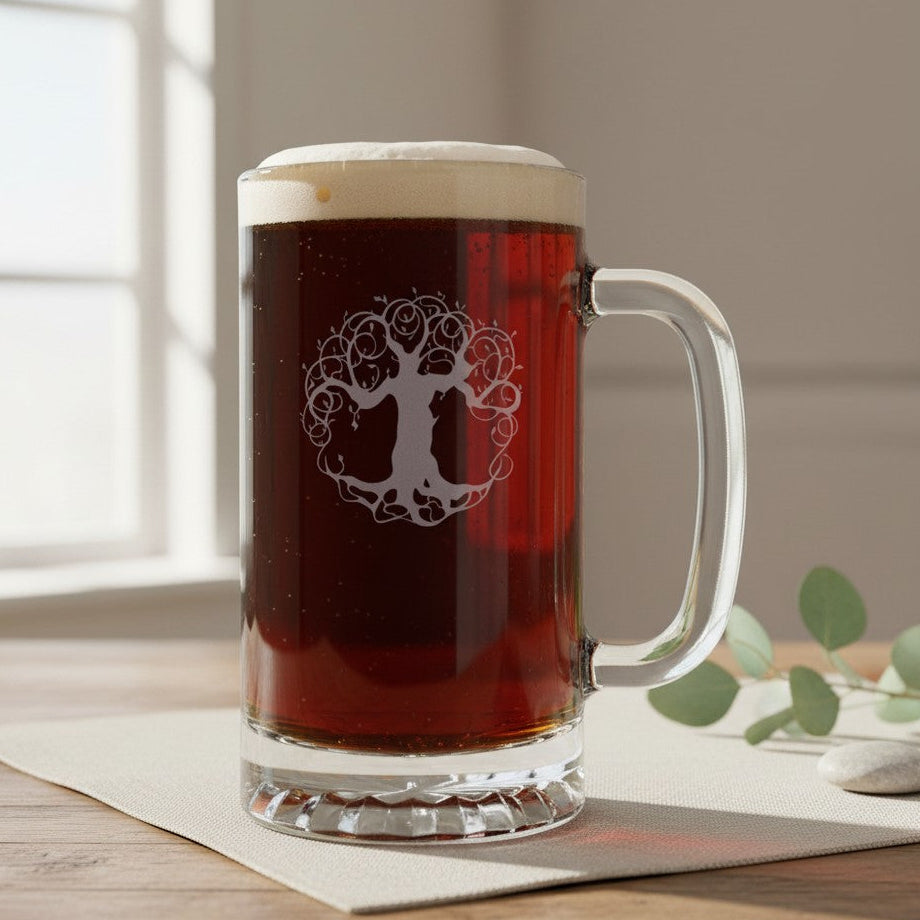 A clear glass beer mug with a handle, filled with dark amber beer and topped with white foam, featuring a white tree-of-life graphic on the front. The mug sits on a beige cloth on a wooden table with soft window light in the background.