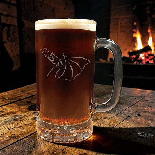 A large clear glass beer mug filled with amber beer and topped with white foam, featuring an etched winged Dragon design on the front, set on a rustic wooden table in front of a warm fireplace.