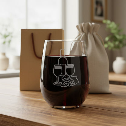 A clear stemless wine glass filled with red wine, featuring an etched wine bottle, two wine glasses, and a grape cluster design on the front. The glass is set on a wooden table with a brown gift bag and a beige drawstring pouch in a softly blurred background.