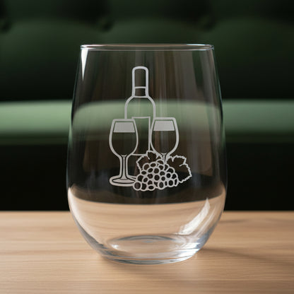 A clear stemless wine glass with a white etched graphic of a wine bottle, two wine glasses, and a grape cluster, shown on a wooden surface against a green couch.