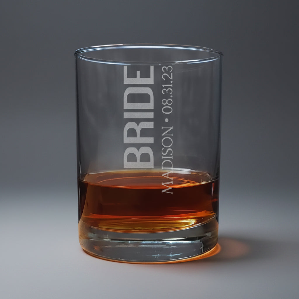 Clear glass tumbler with 'BRIDE' and personalized text filled with amber liquid on a wooden surface.