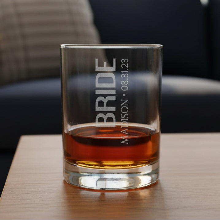 Clear glass tumbler with 'BRIDE' and personalized text filled with amber liquid on a wooden surface.