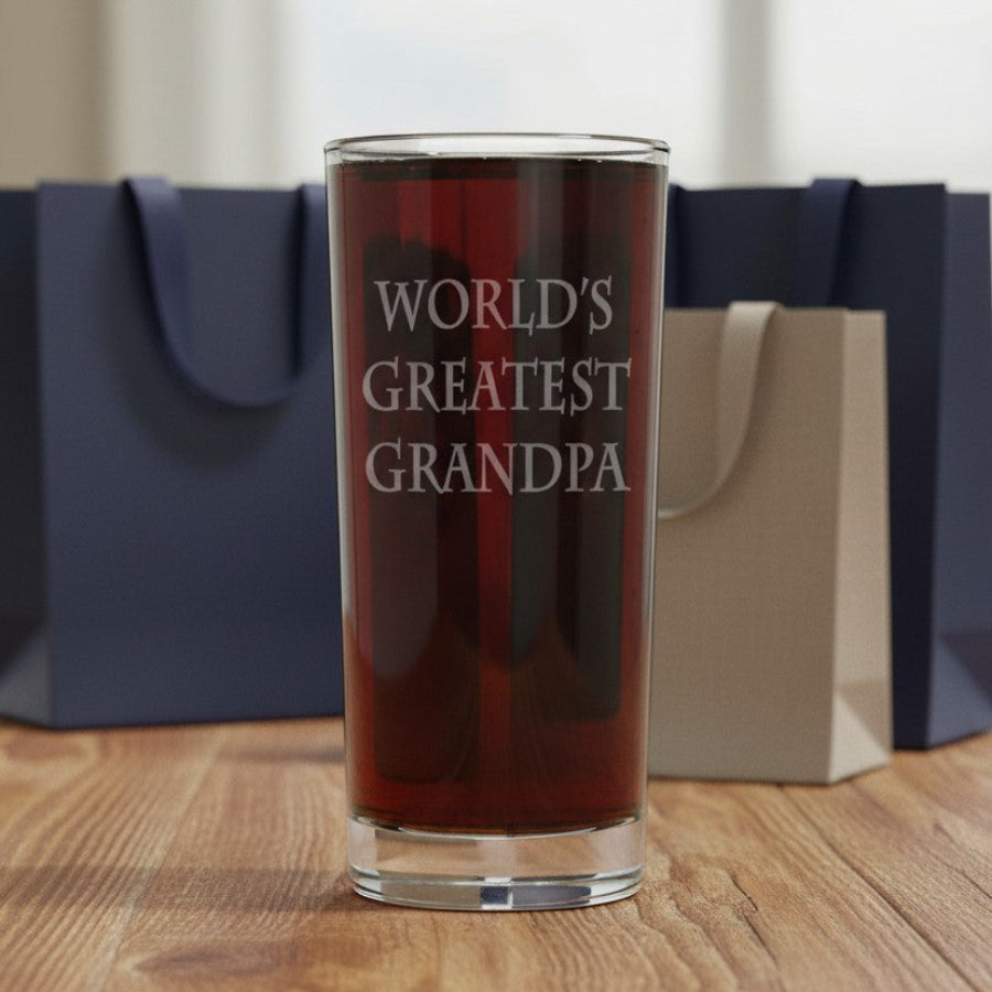 A clear pint glass filled with dark red liquid stands on a wooden table, etched with the phrase 'WORLD'S GREATEST GRANDPA' . Behind it are two gift bags, one navy blue and one beige, against a softly blurred indoor background.
