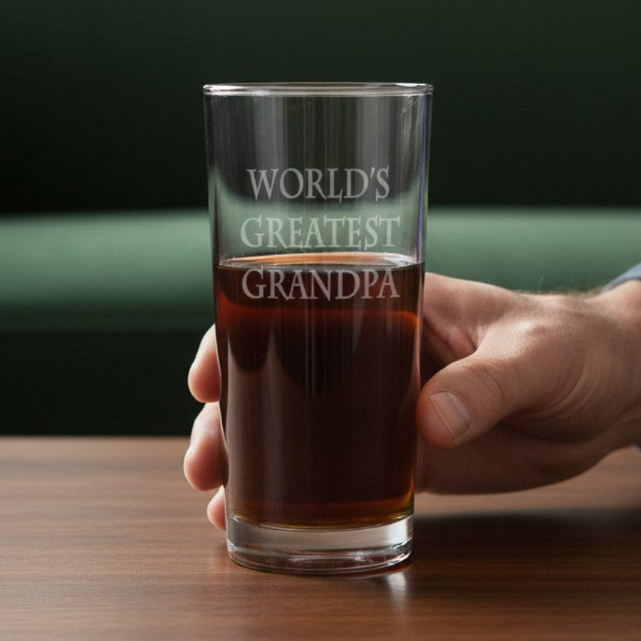 A hand holding a clear glass pint with the phrase 'WORLD'S GREATEST GRANDPA' etched on it, filled with an amber drink, set on a wooden table in front of a green sofa.