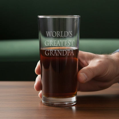 A hand holding a clear glass pint with the phrase 'WORLD'S GREATEST GRANDPA' etched on it, filled with an amber drink, set on a wooden table in front of a green sofa.