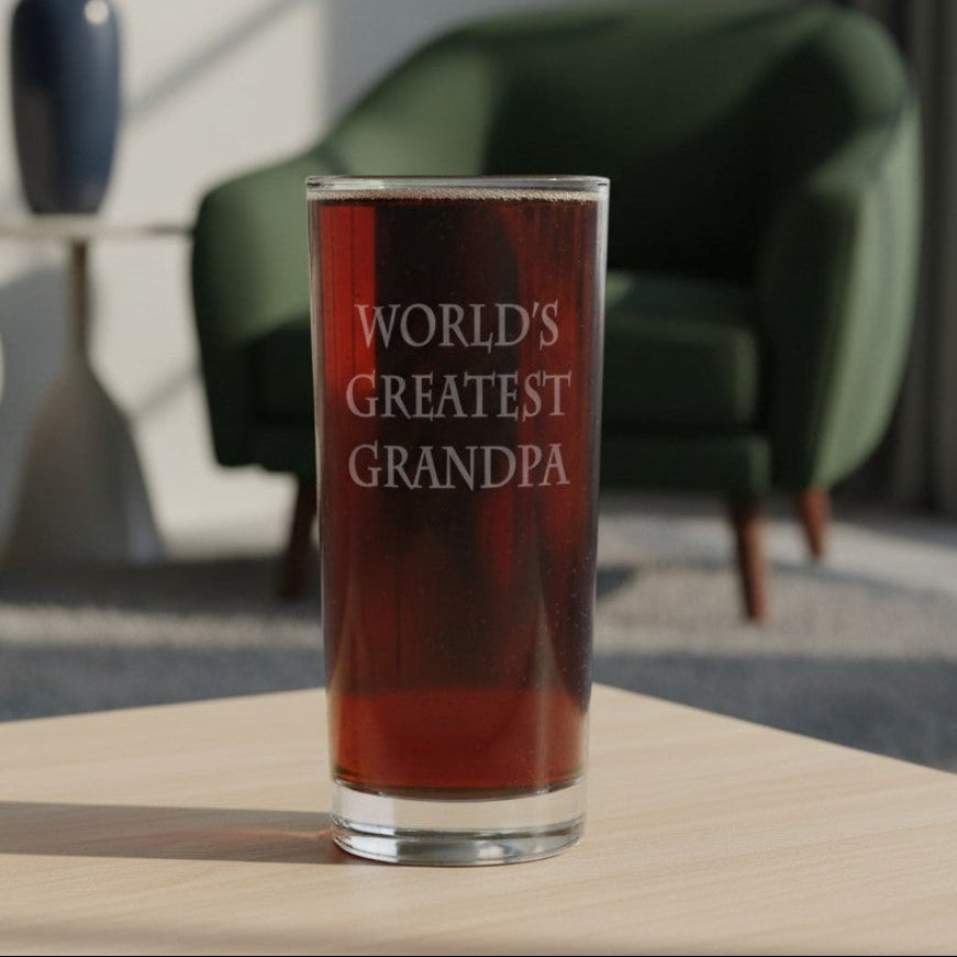 A clear highball glass filled with a dark reddish-brown liquid, standing on a light wood table in a softly blurred living room. The glass is etched with “WORLD’S GREATEST GRANDPA” 