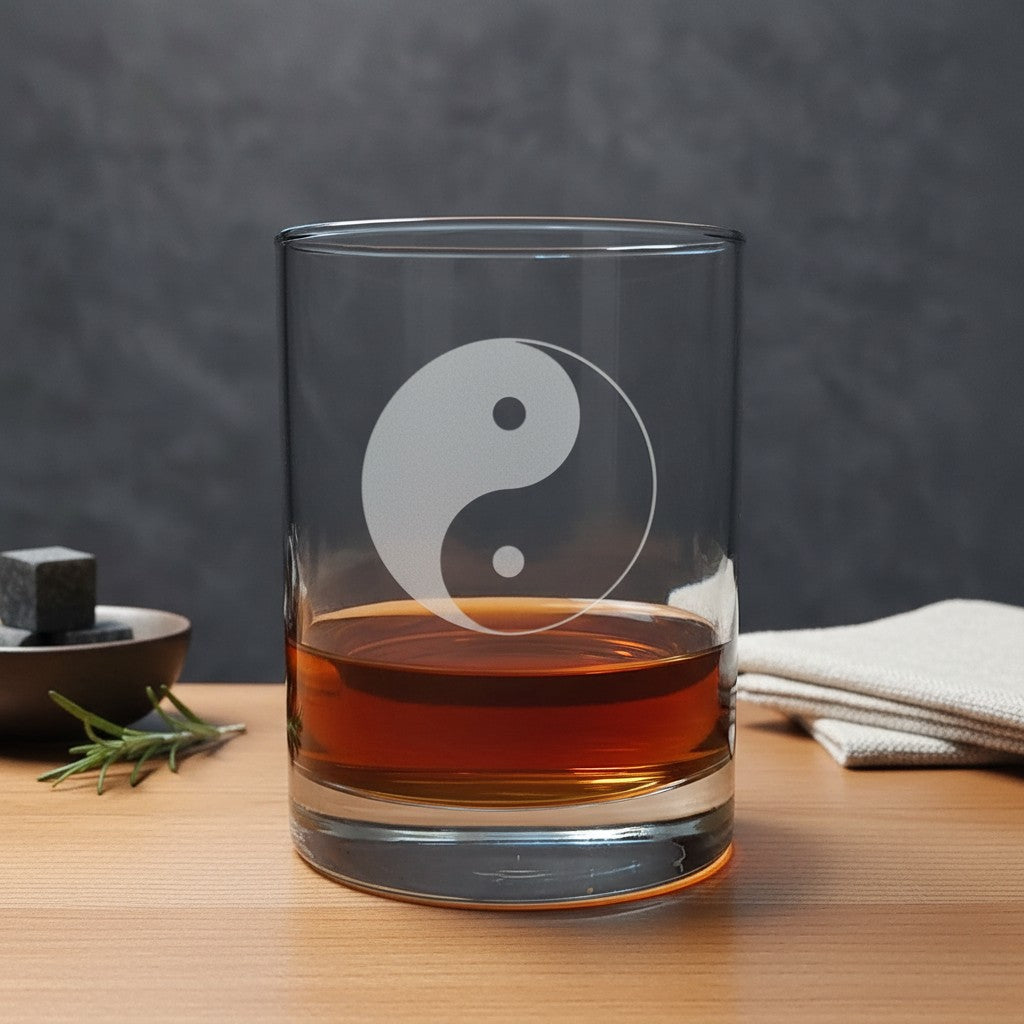 A clear glass rocks tumbler with an etched white yin-yang symbol, partially filled with amber whiskey, set on a wooden table with a dark gray textured background.