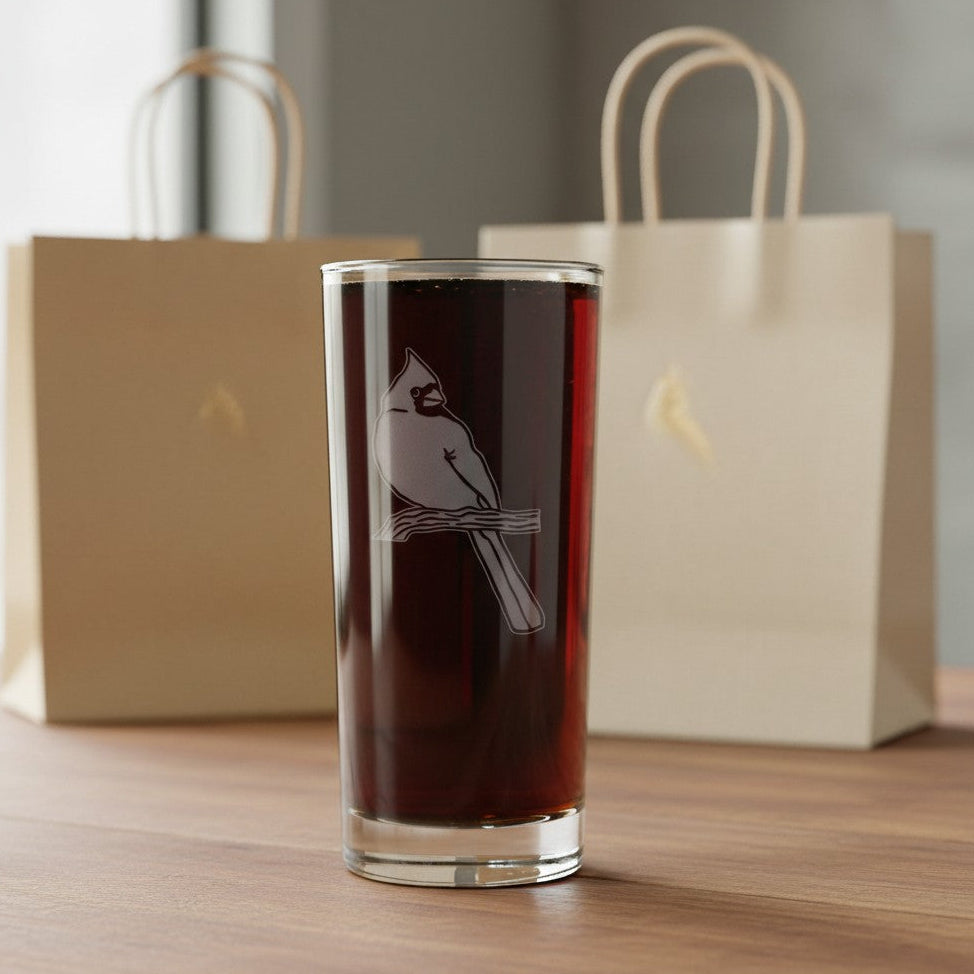 A clear glass tumbler with an etched cardinal perched on a branch, filled with a dark reddish-brown liquid, displayed on a wooden table with two beige gift bags in the background.