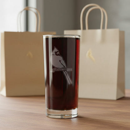 A clear glass tumbler with an etched cardinal perched on a branch, filled with a dark reddish-brown liquid, displayed on a wooden table with two beige gift bags in the background.