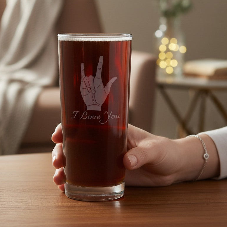 A hand holds a clear glass tumbler filled with a dark reddish-brown beverage on a wooden table. The glass is etched with an etched ASL hand gesture and the words 'I Love You'.