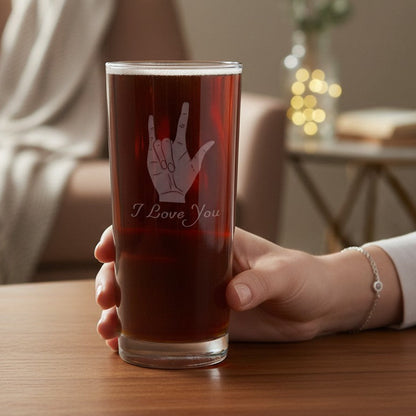 A hand holds a clear glass tumbler filled with a dark reddish-brown beverage on a wooden table. The glass is etched with an etched ASL hand gesture and the words 'I Love You'.