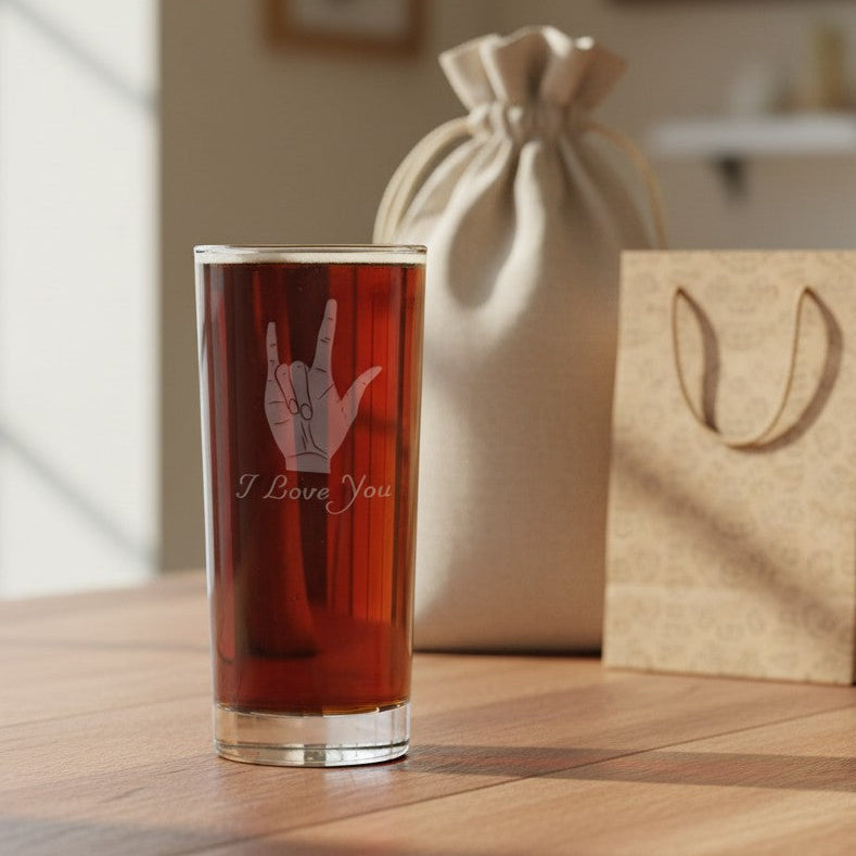 A clear glass tumbler filled with amber liquid stands on a wooden table, etched with a ASL hand gesture and the words 'I Love You.' Behind it are a beige drawstring pouch and a kraft paper gift bag in a softly lit indoor setting.