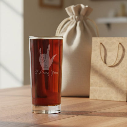 A clear glass tumbler filled with amber liquid stands on a wooden table, etched with a ASL hand gesture and the words 'I Love You.' Behind it are a beige drawstring pouch and a kraft paper gift bag in a softly lit indoor setting.